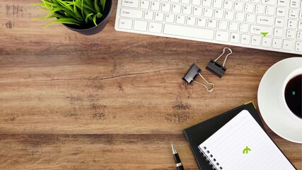 Overhead view of a wooden desk with a white keyboard a potted plant two binder clips a notebook and a coffee cup with dollar signs symbolizing financial success and business productivity