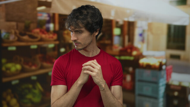 Man in red shirt clasping hands and glancing sideways at a street fruit market stall; quiet contemplation.