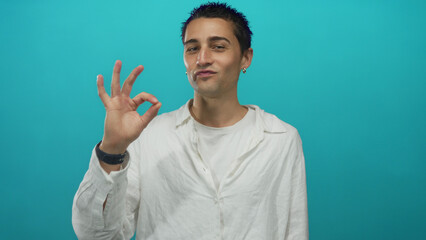 Young man in white shirt making okay gesture against vibrant blue wall expressing positive emotion...