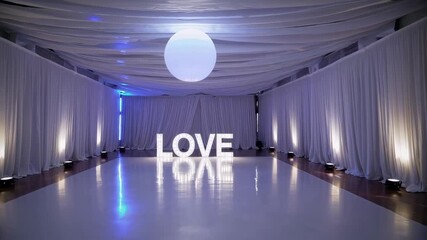Elegant white wedding reception venue with illuminated love sign on dance floor and draped curtains, romantic celebration atmosphere