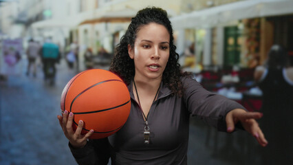 Woman holding basketball talking on outdoor restaurant terrace depicting sports in urban street setting with casual attire and dynamic gestures.