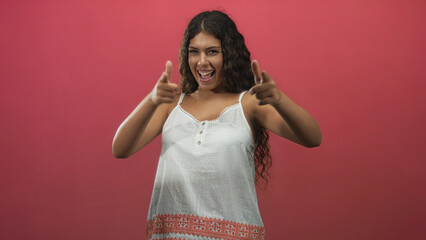 Hispanic woman points finger guns with visible hands and smiling face in a white sundress against a pink backdrop in studio; playful confidence.