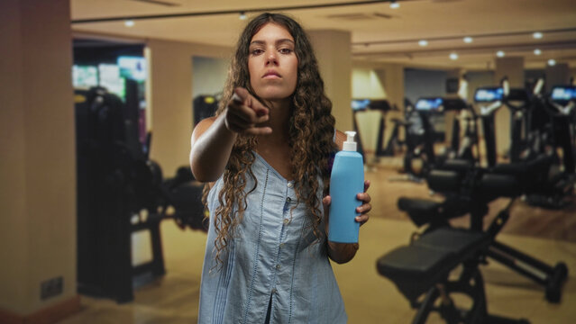 Woman holding blue pump bottle, wearing sleeveless light blue dress, points finger toward camera in gym with cardio machines visible; warning hygiene care.