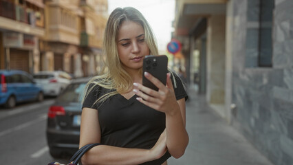 Obraz premium Woman tapping smartphone on street, carrying handbag with fingers on screen, wearing black v neck shirt amid parked car and stone building; concentration communication.
