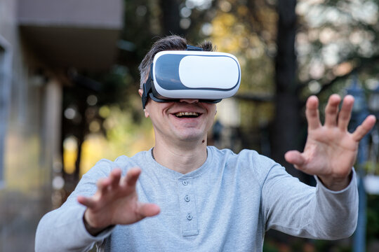 Happy middle-aged man smiling and gesturing with hands while wearing a virtual reality headset outdoors, engaging with a simulation