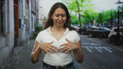 Woman gesturing with hands on a city street wearing a white ribbed top and blue jeans near parked cars and leafy trees; awkward conversation embarrassment.