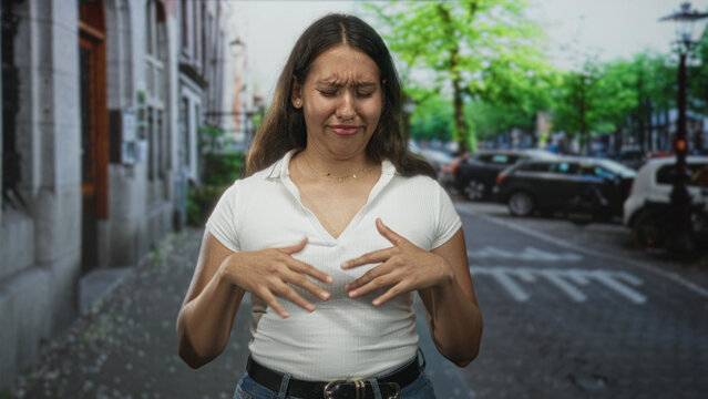 Woman gesturing with hands on a city street wearing a white ribbed top and blue jeans near parked cars and leafy trees; awkward conversation embarrassment.