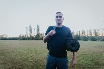 Fototapeta premium Man carrying rolled exercise mat and backpack in open air autumn park after training