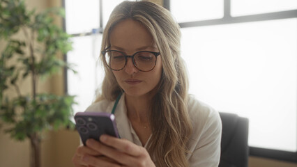 Woman doctor with blonde hair using phone in a clinic office setting, wearing stethoscope and glasses, indoors in modern healthcare workplace.