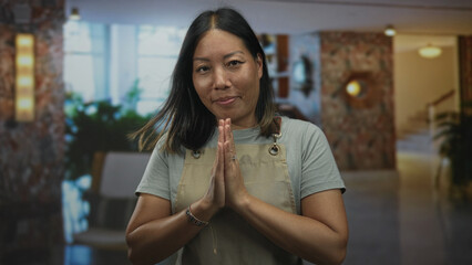 Woman in apron with hands pressed together in building lobby offering a gentle greeting to guests;...