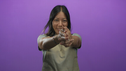 Woman wearing apron clasping hands and extending them toward camera in purple studio; friendly...