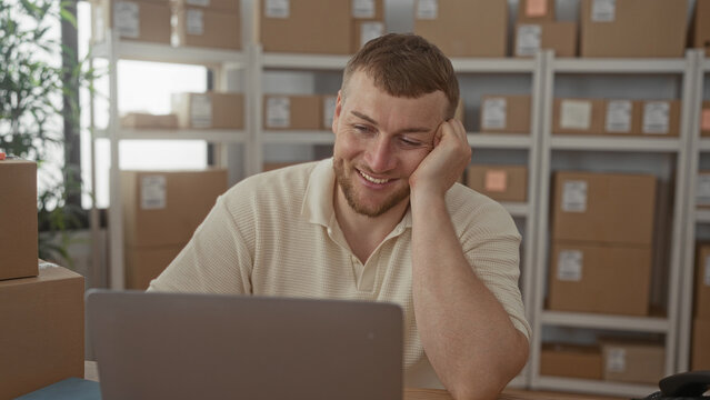 Man smiling and typing at a laptop amid stacked parcels, shipping labels and shelving in a small office building; satisfaction.