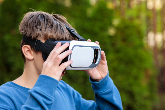 Young teenage boy adjusting and wearing a white and black virtual reality headset outdoors with green foliage background