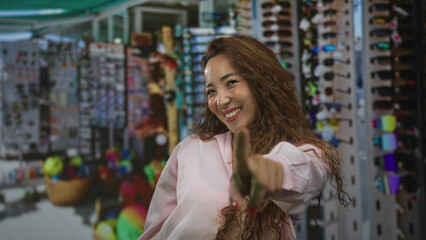 Woman points finger to camera while smiling in a crowded street market souvenir stall with sunglasses rack and baskets visible; playful confidence.