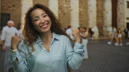 Woman smiling, points finger to camera and hand on chest on old town street; joy confidence warmth.