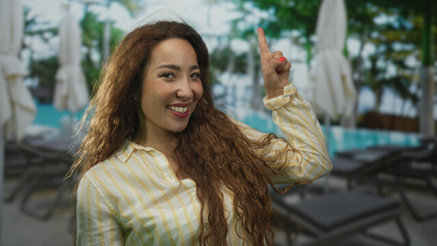 Woman smiling and pointing finger at hotel poolside lounge chairs and umbrellas wearing yellow striped shirt with long curly hair; vacation joy.