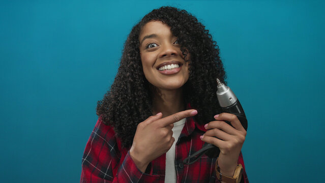 African american woman with curly hair holding drill smiles while pointing finger to drill in blue studio; joy. - Powered by Adobe
