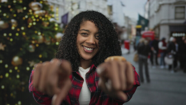 Woman in red plaid shirt points finger at camera on busy city street by christmas tree; friendly invitation.