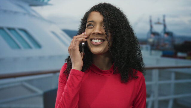 African american woman holding smartphone to ear and smiling while standing on a cruise boat deck under cloudy sky; joy. - Powered by Adobe
