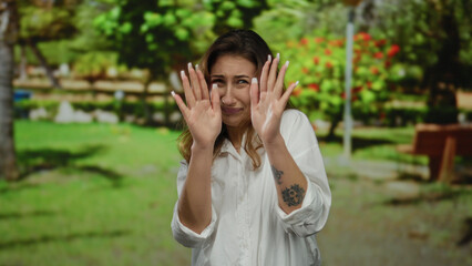 Fototapeta premium Young woman looking fearful with hands covering face in vibrant park setting surrounded by greenery and flowers, expressing emotion outdoors.