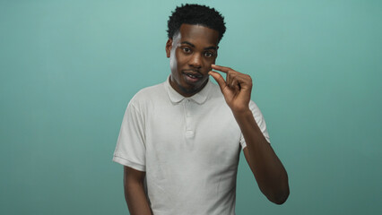 Young african american man smiles and pinches fingers to show a tiny gap in teal studio;...