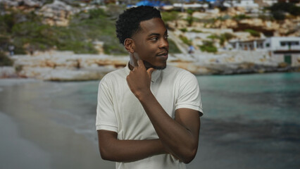 Man in white polo shirt touches cheek with finger on rocky seaside shore with calm water lapping at sand; contemplation.