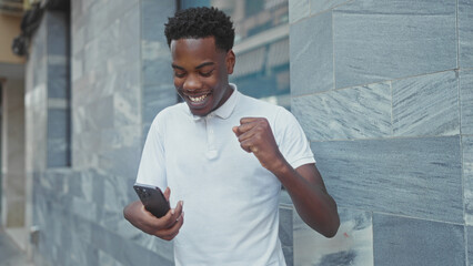 Man leaning against a tiled stone wall on a street holding smartphone lifts clenched fist in victory; excitement triumph victory celebration.