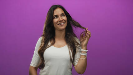 Obraz premium Young hispanic woman smiling, touching hair and wrist bracelets in purple studio setting; playfulness.