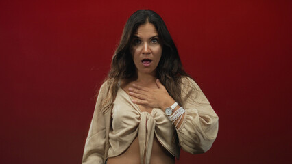 Woman with hand on chest, open mouth and tie front top in studio against deep red backdrop; surprise vulnerability.