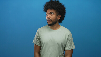 Man with glasses and afro hairstyle shows fear against blue wall background creating isolated and...