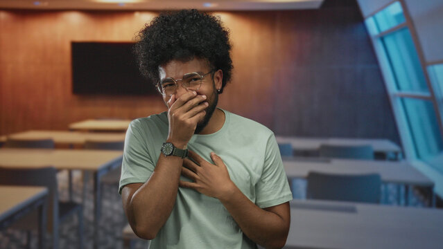 Man laughing in a classroom with hands covering mouth and tables in the background, expressing joy and emotion in an indoor educational setting.