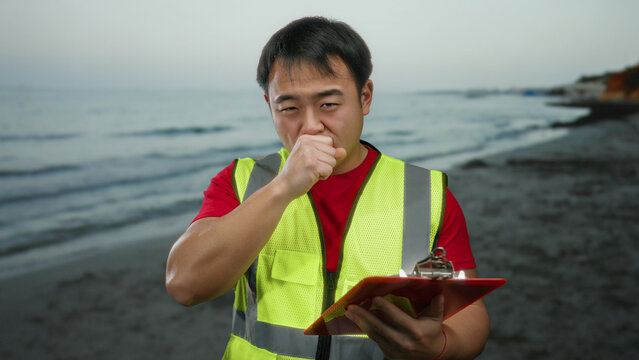 Man wearing a safety vest stands on a sandy beach by the sea, holding a clipboard and coughing into his hand, suggesting outdoor work or supervision. - Powered by Adobe