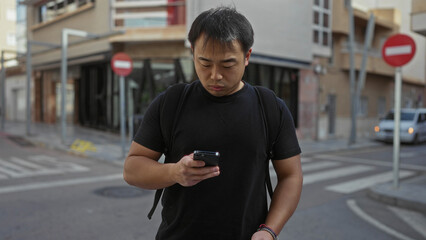 Young man with backpack checking phone on urban street in daytime, showcasing a casual modern lifestyle in city environment.