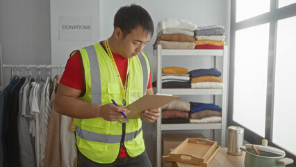 Young man in charity center wearing a safety vest and holding a clipboard, organizing donations in a room filled with clothes and supplies.