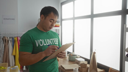 Asian man volunteering in a charity center, wearing a green shirt, taking notes on a clipboard in a room full of donations.