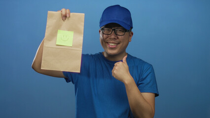 Delivery man holding paper bag with smile note and pointing finger in blue studio; cheerful service.