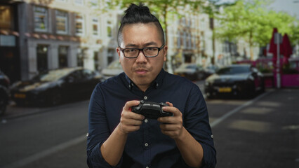 Man holding game controller in both hands, squinting with closed eyes and tense mouth while standing on a busy city street lined with cars and storefronts; frustration.