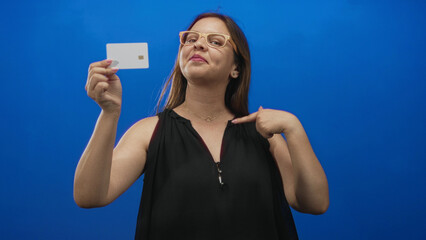 Woman holding a white creditcard and finger pointing to her chest in blue studio; confidence empowerment.