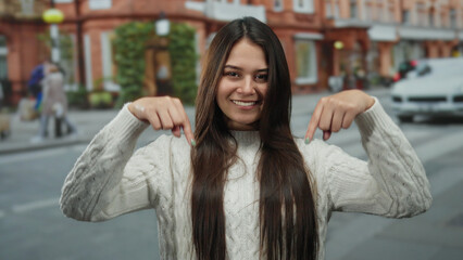 Woman pointing downward stands on a bustling street with historic red brick buildings, exuding confidence and youthful energy in an outdoor urban setting.