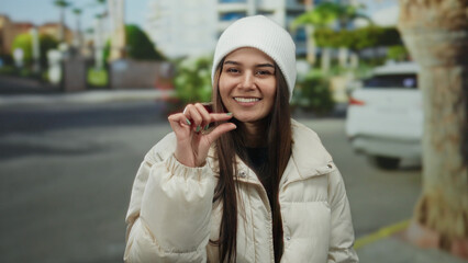 Obraz premium Woman smiling in winter jacket on street, gesturing small size with hand against blurred urban backdrop, showcasing her warm clothing and friendly demeanor outdoors during daytime.
