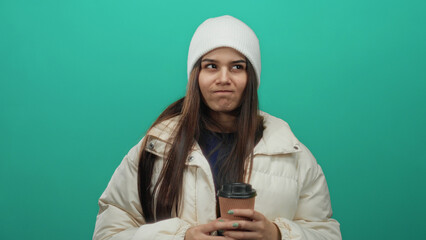 Woman in white jacket and beanie holding coffee cup against green background looks thoughtful, capturing a moment of contemplation with casual winter attire.
