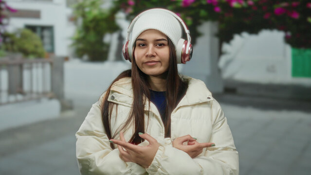 Young woman wearing headphones and a winter jacket listens to music on a peaceful street lined with flowering trees, enjoying a moment outdoors.