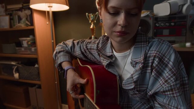 Handheld shot of young female Caucasian zoomer practicing acoustic guitar while sitting at home