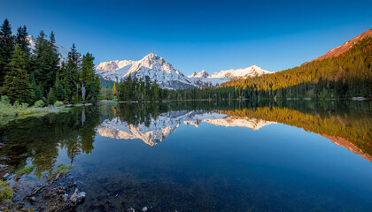 Fototapeta premium Snow Capped Mountains Reflect Beautifully In A Calm Alpine Lake Surrounded By Evergreen Trees Under A Clear Blue Sky At Sunset