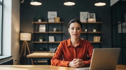 Woman in orange shirt at desk with laptop in office with shelves and lamp behind her - Powered by Adobe