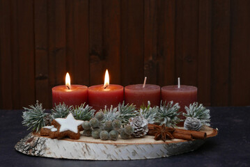 Zweiter Advent: Längliches Adventsgesteck mit braunen Kerzen, Zimtsternen, Zapfen, Beeren, Zimtstange und Tannenzweigen vor einer braunen Holzwand mit Textfreiraum.