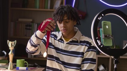 Medium shot of gen Z athletic boy talking about medals and trophies on camera of smartphone in ring light holder while streaming at home - Powered by Adobe