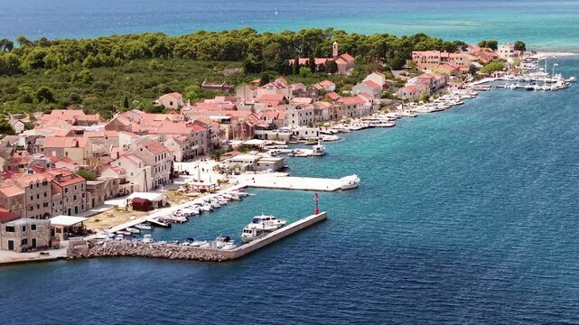 Vibrant coastal scenery of a historic Krapanj village with a busy waterfront promenade, docked yachts, and a ferry boat navigating the calm turquoise waters of the Dalmatian coast