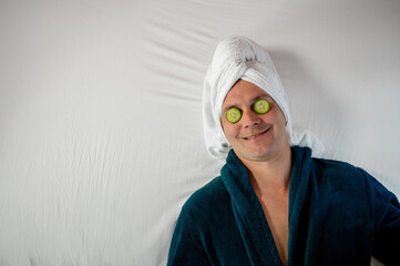 Man with cucumber eye mask in bathrobe, having fun during skincare on white bed, copy space.
