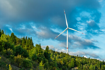 Modern wind turbine generating renewable energy against blue sky with white clouds above lush green forest landscape in countryside